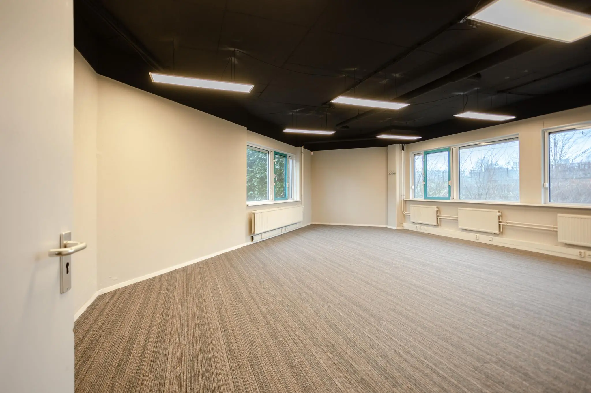 Leeg kantoor met tapijtvloer, zwarte systeemplafonds en grote ramen aan de Christiaan Krammlaan.